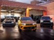A gold Dodge Hornet PHEV parked in front of a mechanic's shop flanked by two other muscle cars.