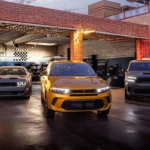 A gold Dodge Hornet PHEV parked in front of a mechanic's shop flanked by two other muscle cars.