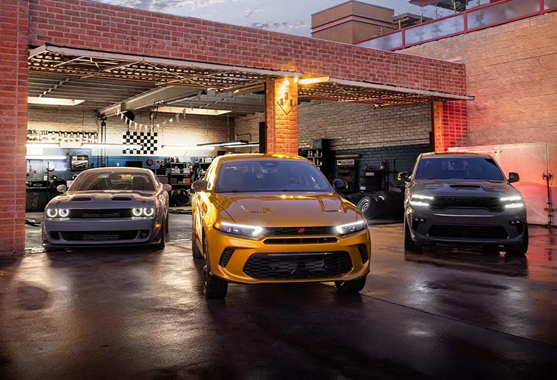 A gold Dodge Hornet PHEV parked in front of a mechanic's shop flanked by two other muscle cars.